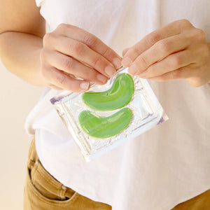 Person holding green eye masks in a clear packaging against a neutral background