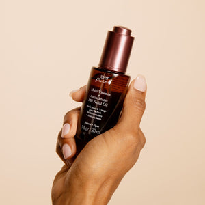 Hand holding a bottle of 100% Pure Multi-Vitamin Antioxidants PM Facial Oil against a beige background