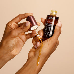 Hand holding a dropper with facial oil and a bottle of facial oil against a beige background
