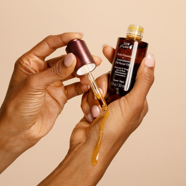 Hand holding a dropper with facial oil and a bottle of facial oil against a beige background