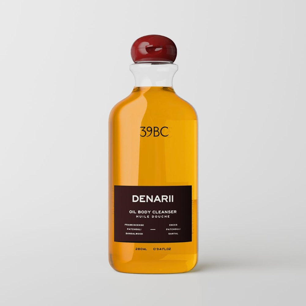 A 280ml glass bottle of DENARII 39BC Oil Body Cleanser, filled with amber-colored liquid and featuring a black label with a dark red stopper, set against a plain white background.