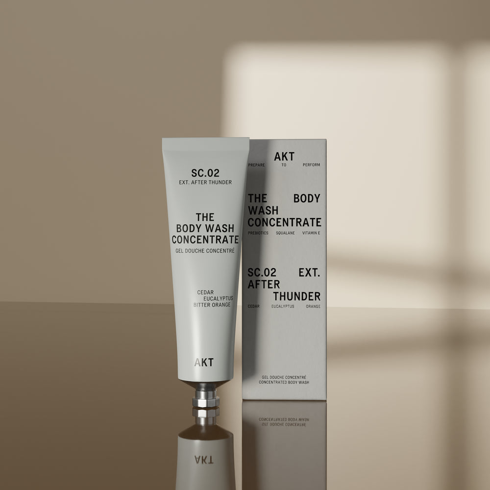 A white squeeze tube of AKT London body wash concentrate, SC.02, with black text, shown on a light gray background with a soft shadow.