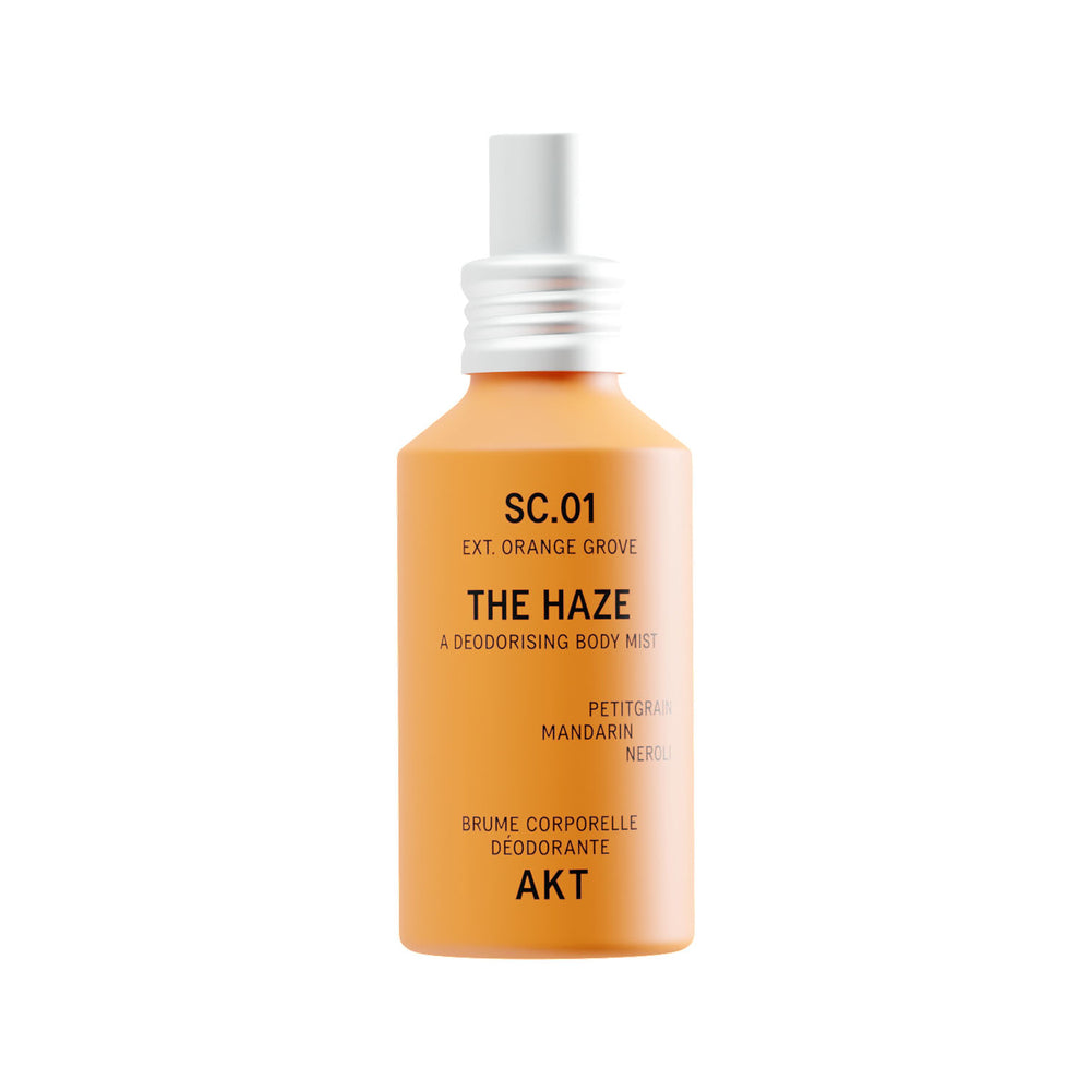 A bright orange body mist bottle with a silver spray nozzle, labeled 'THE HAZE' by the brand AKT London, on a solid white background.