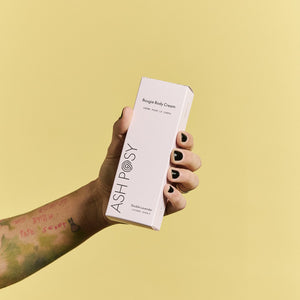 Hand holding a box of Ash Posy body cream against a yellow background