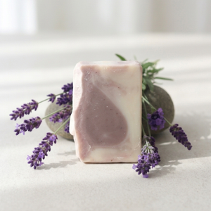 A rectangular bar of lavender soap with a swirled light purple and pale pink pattern, styled with fresh lavender sprigs and a dark stone on a light beige surface.