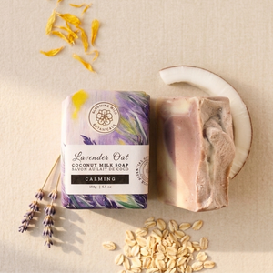 A bar of lavender oat coconut milk soap by Blooming Wild Botanicals, shown wrapped in a purple and green patterned paper sleeve next to an unwrapped bar of the same soap, styled with dried lavender, oats, and a slice of coconut on a beige fabric background.