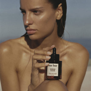 Woman holding a bottle of BUR BUR Growing Season Burdock Hair Oil product against a neutral background