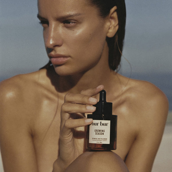 Woman holding a bottle of BUR BUR Growing Season Burdock Hair Oil product against a neutral background