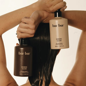 Two bottles of Bur Bur hair care products are held by a woman at the back of her head with wet hair against a neutral background.