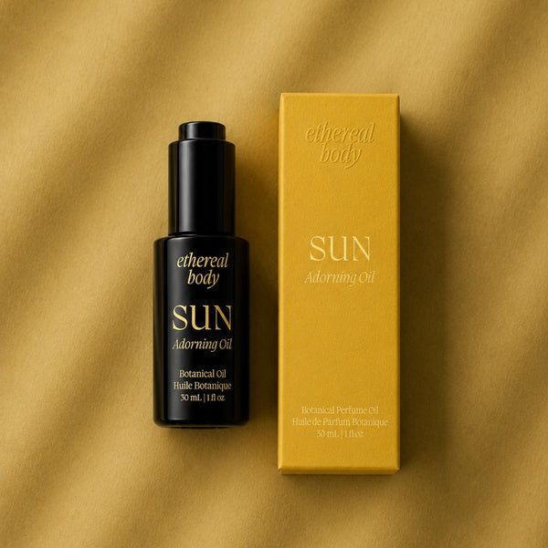 A black bottle of ethereal body SUN Adorning Oil next to its mustard yellow box, set against a textured mustard yellow background.