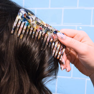 Woman using a colorful comb labeled 'everist' on their hair against a blue tiled wall.