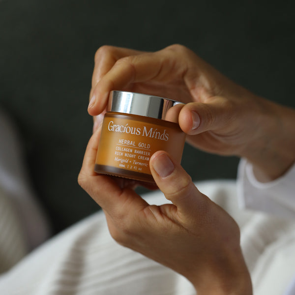 Hands holding a jar of 'Gracious Minds Herbal Gold' cream against a dark background