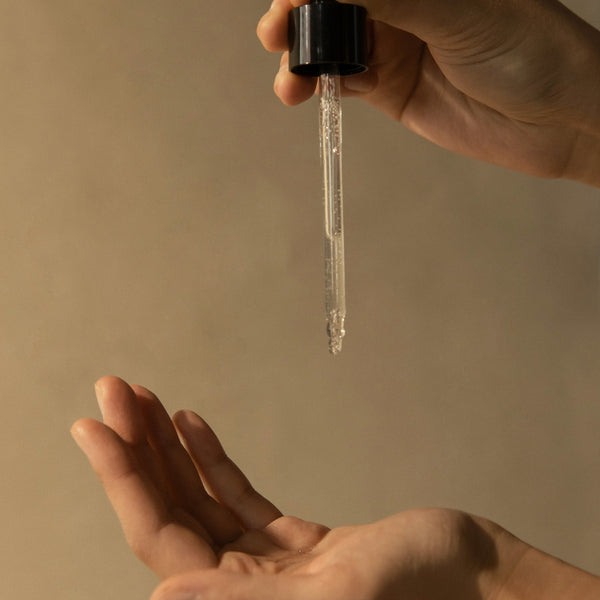 Hand holding a dropper with clear face serum above another hand on a beige background