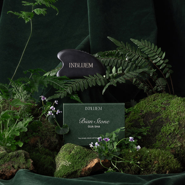 Inbluem Bian Stone Gua Sha tool surrounded by greenery on a dark background