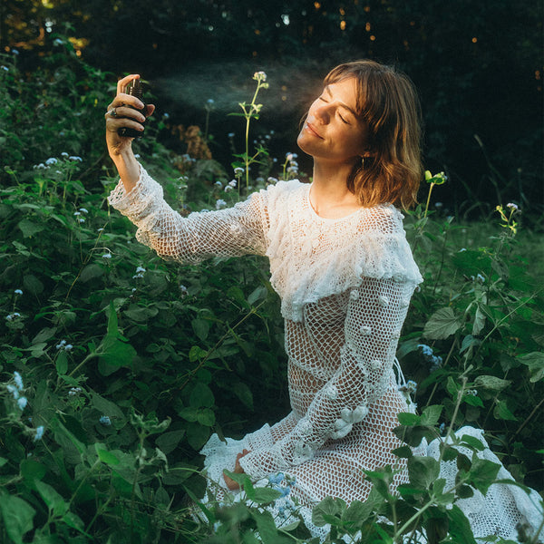 Woman in a white dress sitting in a garden, holding a spray bottle and misting her face. She is using INBLUEM Hydrosoul Cleansing & Toning Mist.