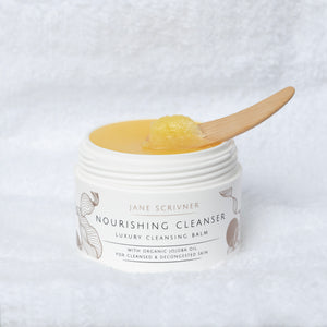 Jane Scrivner Nourishing Cleanser jar with a wooden spoon holding yellow cleansing balm, set against a textured white background.