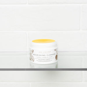 Jane Scrivner Nourishing Cleanser in a white jar with a yellow lid, displayed on a glass shelf against a white subway tile background.