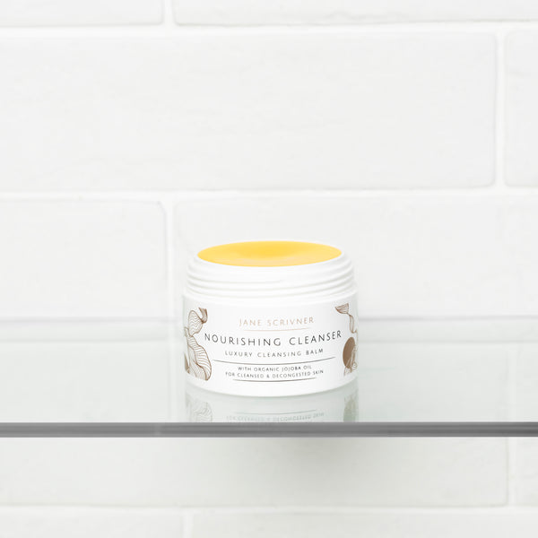Jane Scrivner Nourishing Cleanser in a white jar with a yellow lid, displayed on a glass shelf against a white subway tile background.