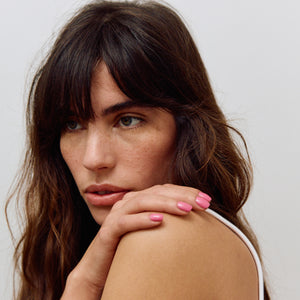 Woman with long brown hair and pink nail polish resting her head on her hand against a plain background