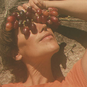 A woman holding red grapes and placing them on her forehead against a textured wall. LEPAAR Summer Wine / Even Tone Antioxidant Shield / Zero Essential Oils ALWAYS SHOW
