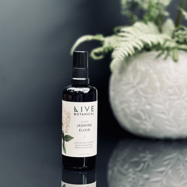 Bottle of Live Botanical Jasmine Elixir on a reflective surface with a blurred plant in the background.