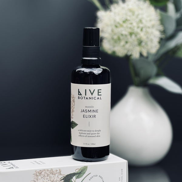Bottle of Live Botanical Jasmine Elixir with a white vase and flowers in the background
