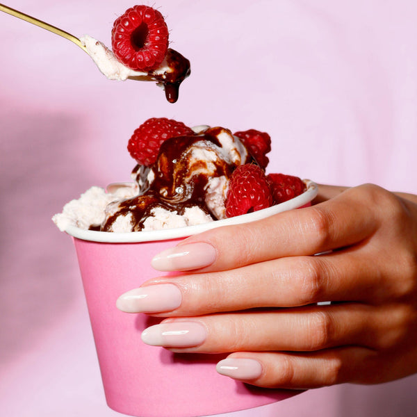 Pink ice cream cup with vanilla ice cream, raspberries, and chocolate sauce held by a hand with long nails and sheer creamy white color against a pink background.