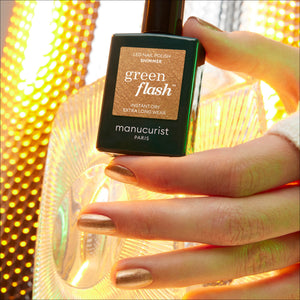 A hand holding a bottle of Manucurist Green Flash LED nail polish in shade Shimmer, showing metallic gold painted fingernails against a warm, glowing gold background.