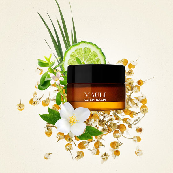 A glass amber jar of MAULI RITUALS Sleep Dharma Calm Balm surrounded by ingredients like chamomiles and a slice of lime on very light beige background.