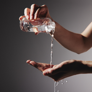 Woman pouring face toner from a clear glass bottle into her other hand against a grey background
