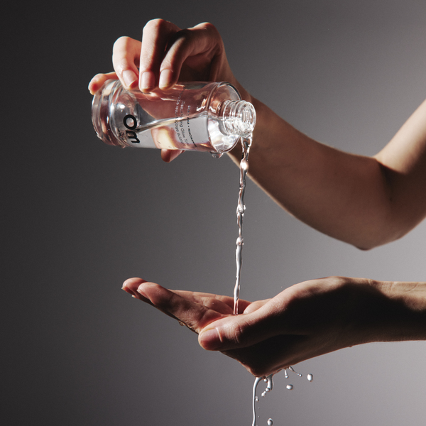 Woman pouring face toner from a clear glass bottle into her other hand against a grey background