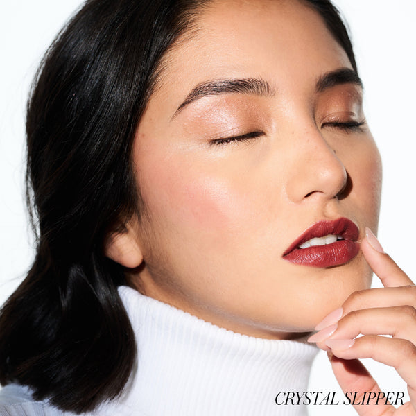 Close-up of a woman with red lipstick and a white turtleneck. Wearing RMS BEAUTY Luminizer Crystal Slipper
