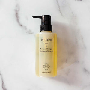 A bottle of Ruhaku Thalasse Shampoo with a black pump dispenser, filled with a pale yellow liquid, set on a white marble background.