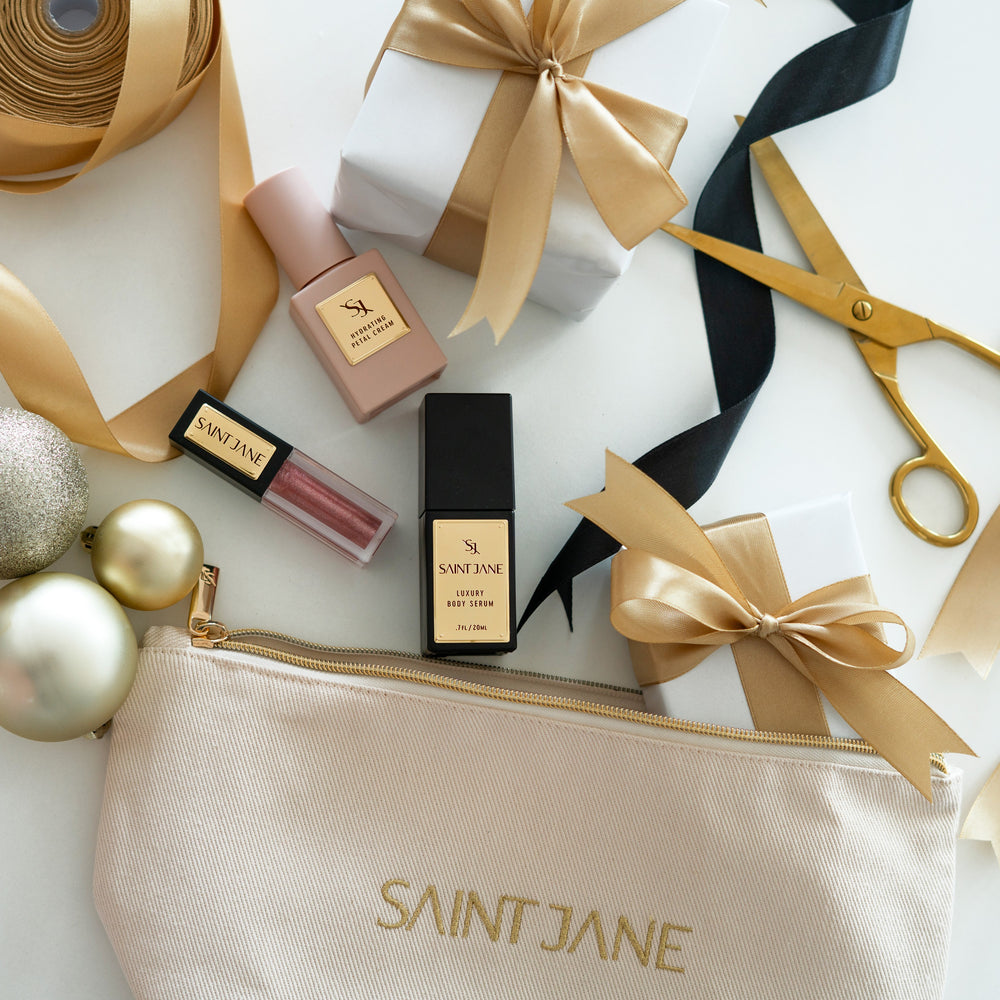 SAINT JANE Little Luxuries Holiday Glow Set with deluxe Hydrating Petal Cream, Luxury Body Serum, Luxury Lip Oil in Alchemy, and a gold makeup bag.