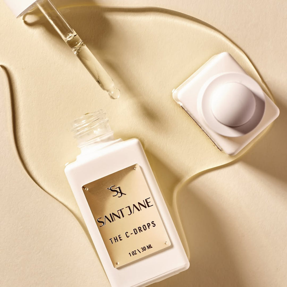 A white glass bottle with white dropper and gold label of SAINT JANE Vitamin C Glow Drops Brightening Serum is on white background