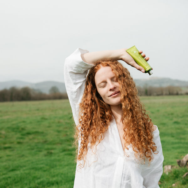 A woman with long, curly red hair wearing a white shirt stands in a green field, holding a green tube above her head. The background shows a soft-focus landscape with hills and trees under an overcast sky.