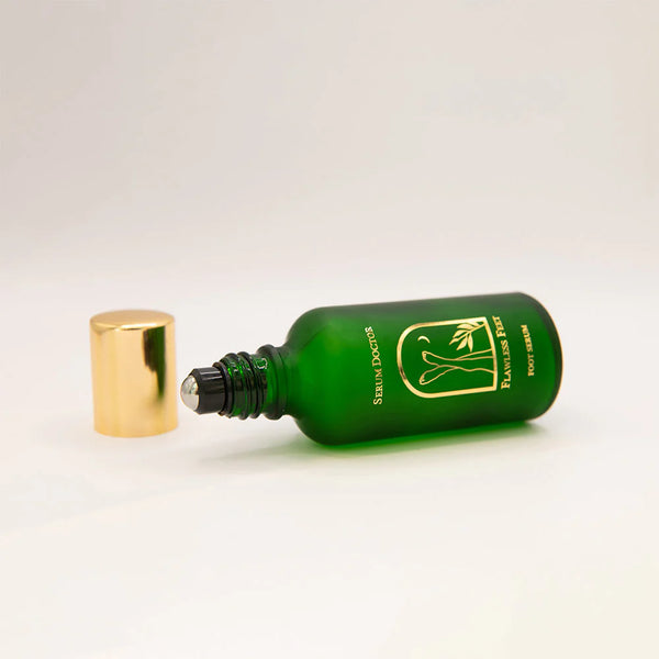 A green glass roll-on bottle with a gold cap lying on its side, next to its detached gold cap, on a clean white background. The bottle features a gold logo and text that reads 'SERUM DOCTOR' and 'Flawless Feet Serum'.