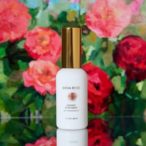 A white bottle of Shiva Rose Radiant Rose Water with a metallic gold cap, set against a blurred background of pink and red roses.