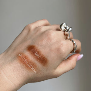 Hand with makeup swatches labeled 'Tan' and 'Golden Medium' on a neutral background
