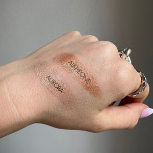 Hand swatching makeup products labeled 'Aurora' and 'Alkheone' on a neutral background