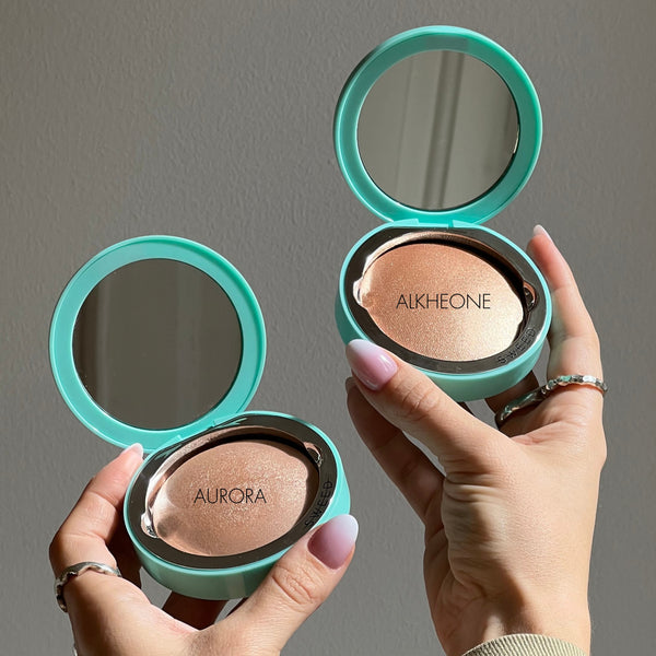 Two turquoise compacts with mirrors, one labeled 'Aurora' and the other 'Alkheone', held by hands against a gray background.