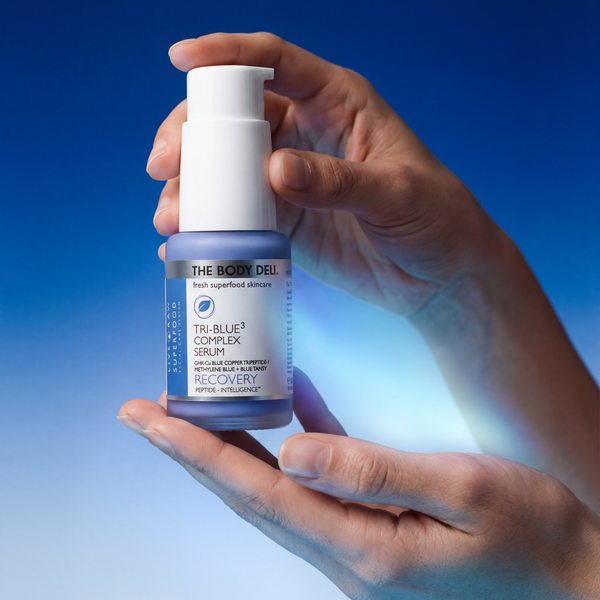 Hand holding a bottle of 'The Body Deli' Tri-Blue Complex Serum against a blue gradient background