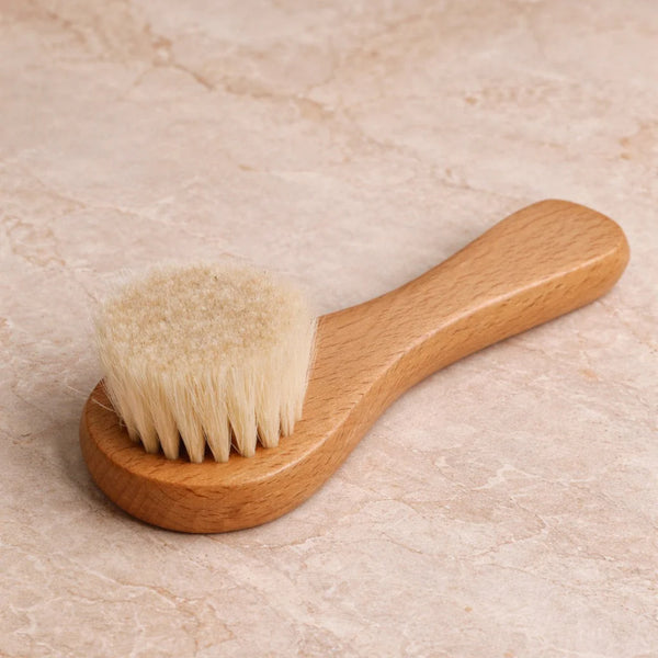 A small wooden face brush with a light natural wood handle and soft cream-colored bristles, resting on a beige stone-textured surface.