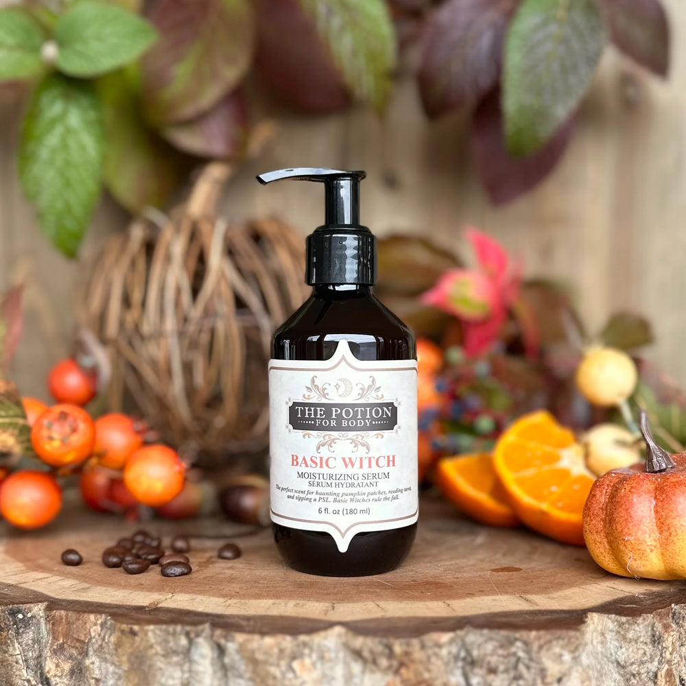 THE POTION COSMETICS Basic Witch Body Potion bottle surrounded by autumn leaves, highlighting pumpkin, coffee, vanilla, and patchouli notes.