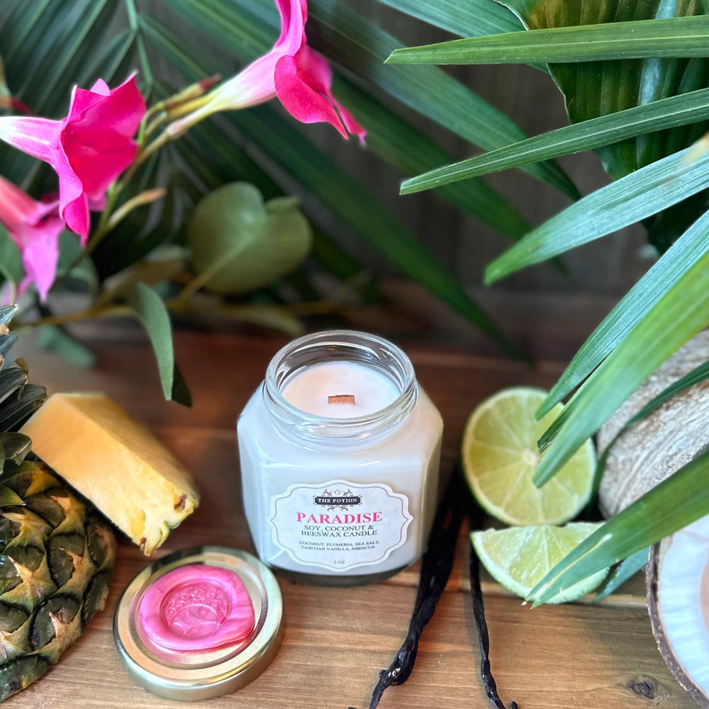 Jar of 'Paradise' candle from The Potion Cosmetics with tropical elements like a pineapple, lime, and flowers in the background.