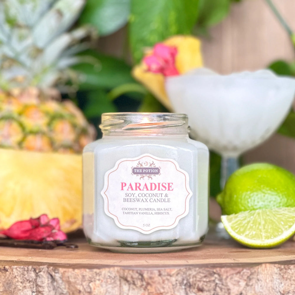 Jar of 'Paradise' candle from The Potion Cosmetics with tropical elements like a pineapple, lime, and flowers in the background.