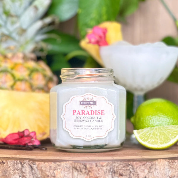 Jar of 'Paradise' candle from The Potion Cosmetics with tropical elements like a pineapple, lime, and flowers in the background.