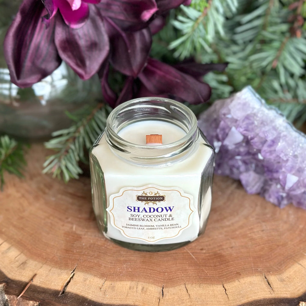 THE POTION COSMETICS Shadow Candle in a glass jar with wooden wick, surrounded by vanilla, jasmine, tobacco leaf, and patchouli, evoking a cozy, uplifting floral aroma.