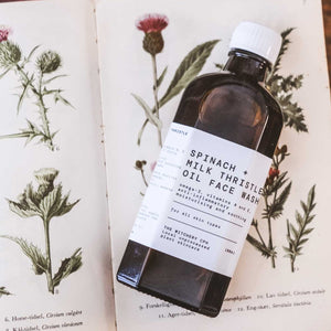 A dark amber glass bottle of Spinach and milk thistle oil face wash with a white cap, resting on an open vintage-style botanical book.