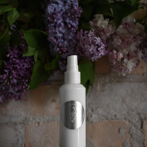 White spray bottle with a silver label on a textured surface with purple flowers in the background. TIDELANDS HOUSE Lilac Nectar Artisanal Copper Distillate